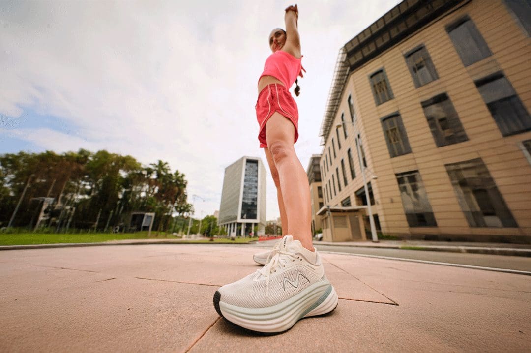 Kvinde i rødt sportstøj strækker ud iført hvide MBT M-3000 II fysiologiske sneakers med tyk rullesål.