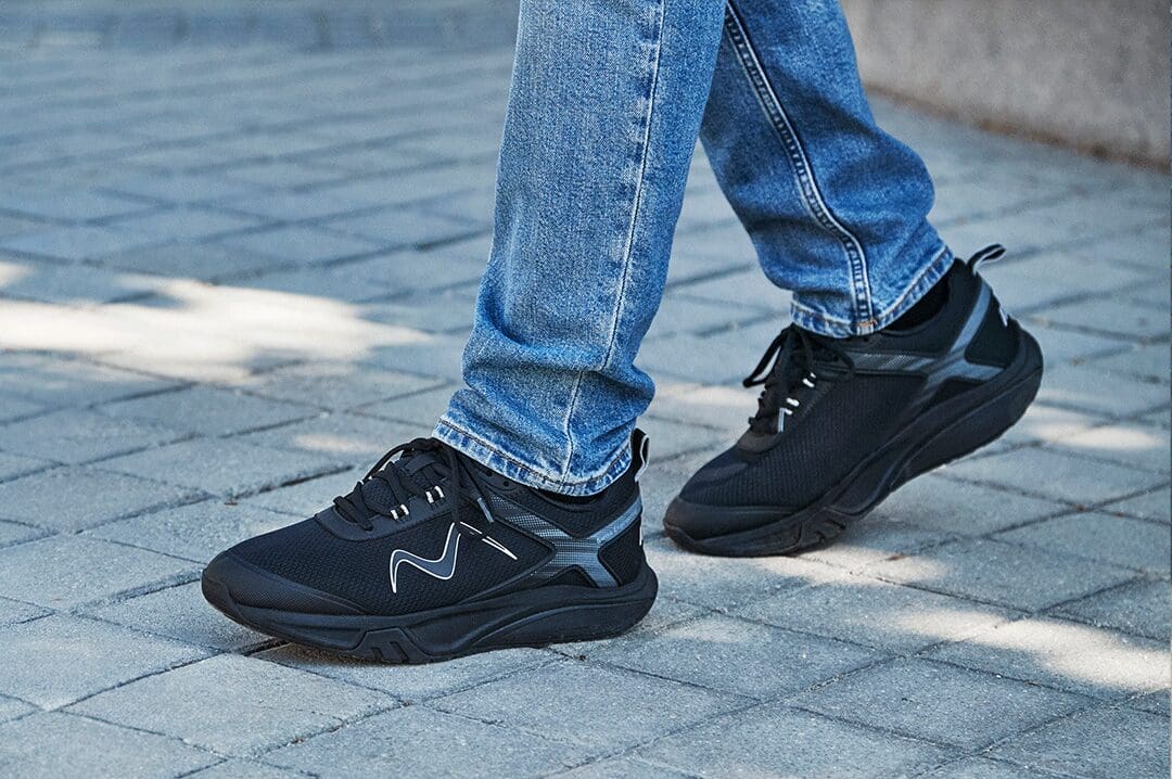 Mand i blå jeans og sorte MBT Sport 4 III herresko med rullesål på flisegang.