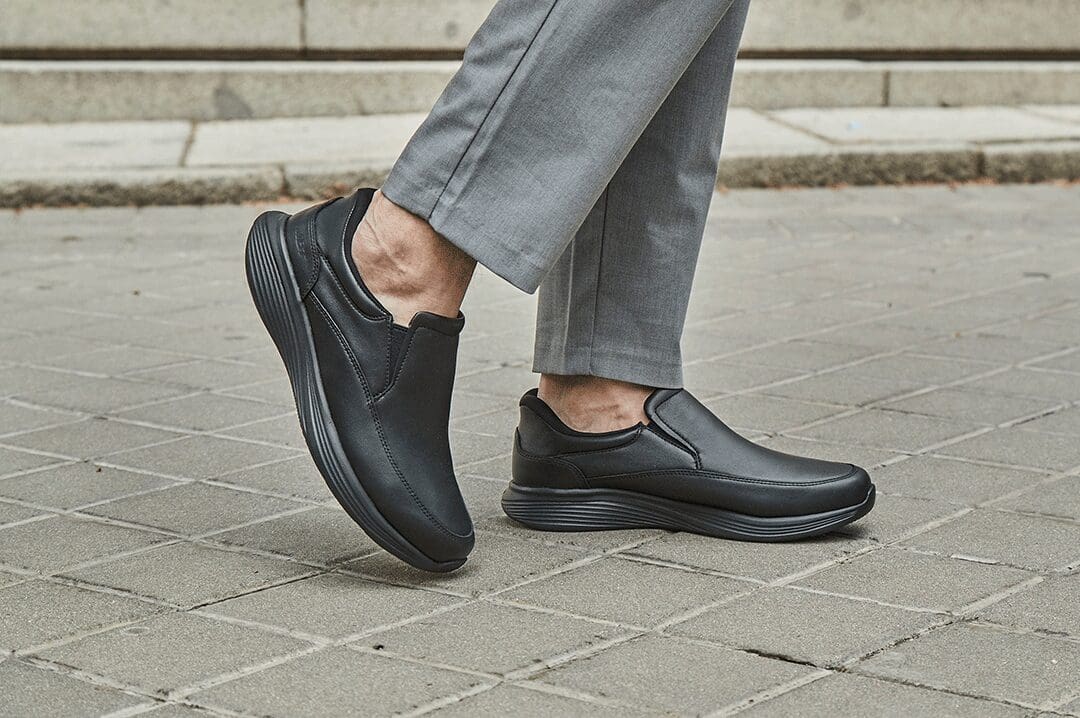 Mand i grå bukser bærer sorte MBT SF-2000 slip-on sko i skind på en flisebelagt vej.