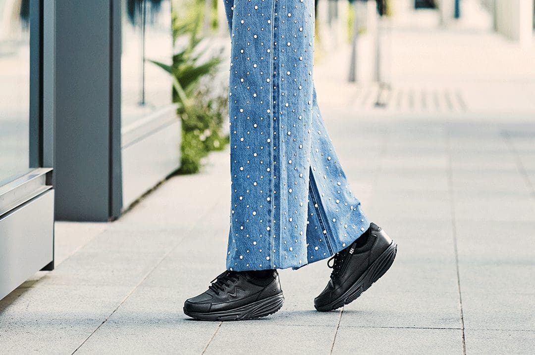 Kvinde i jeans med perler bærer sorte MBT Simba Trainer II sneakers med rullesål på en flisebelagt sti.