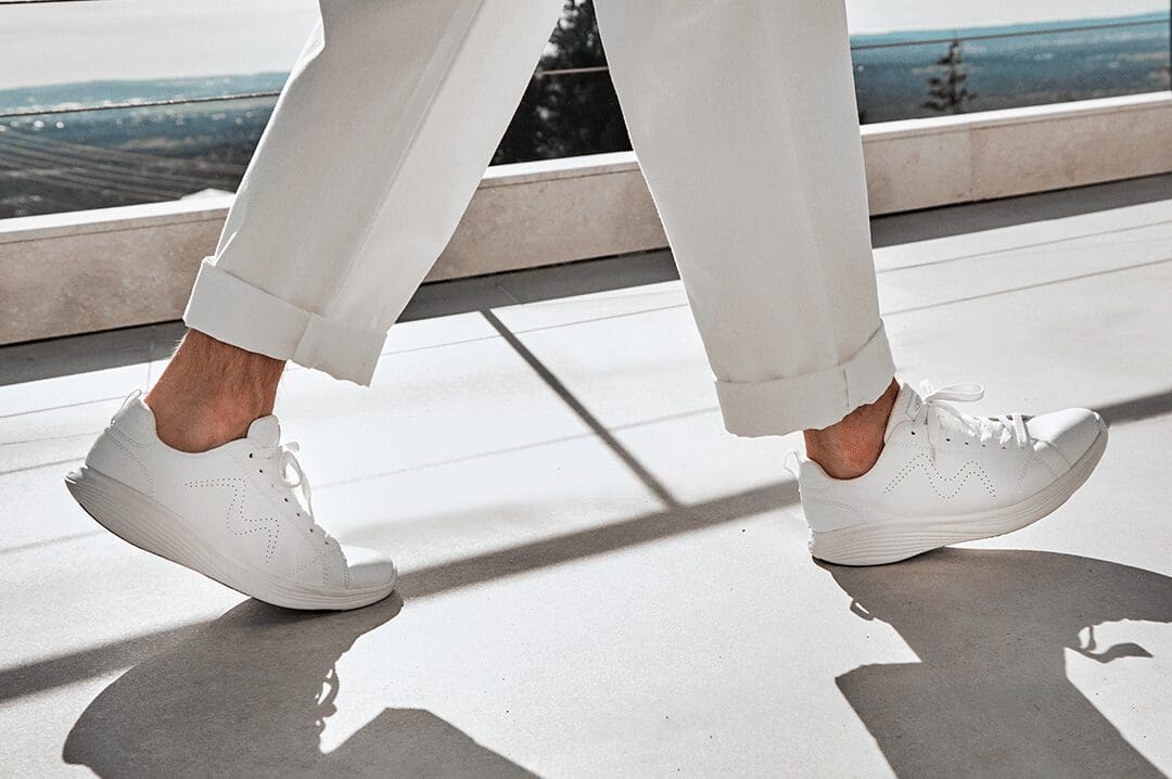 Mand i hvide MBT Ren sneakers og hvide bukser går på en solrig terrasse med smuk udsigt.